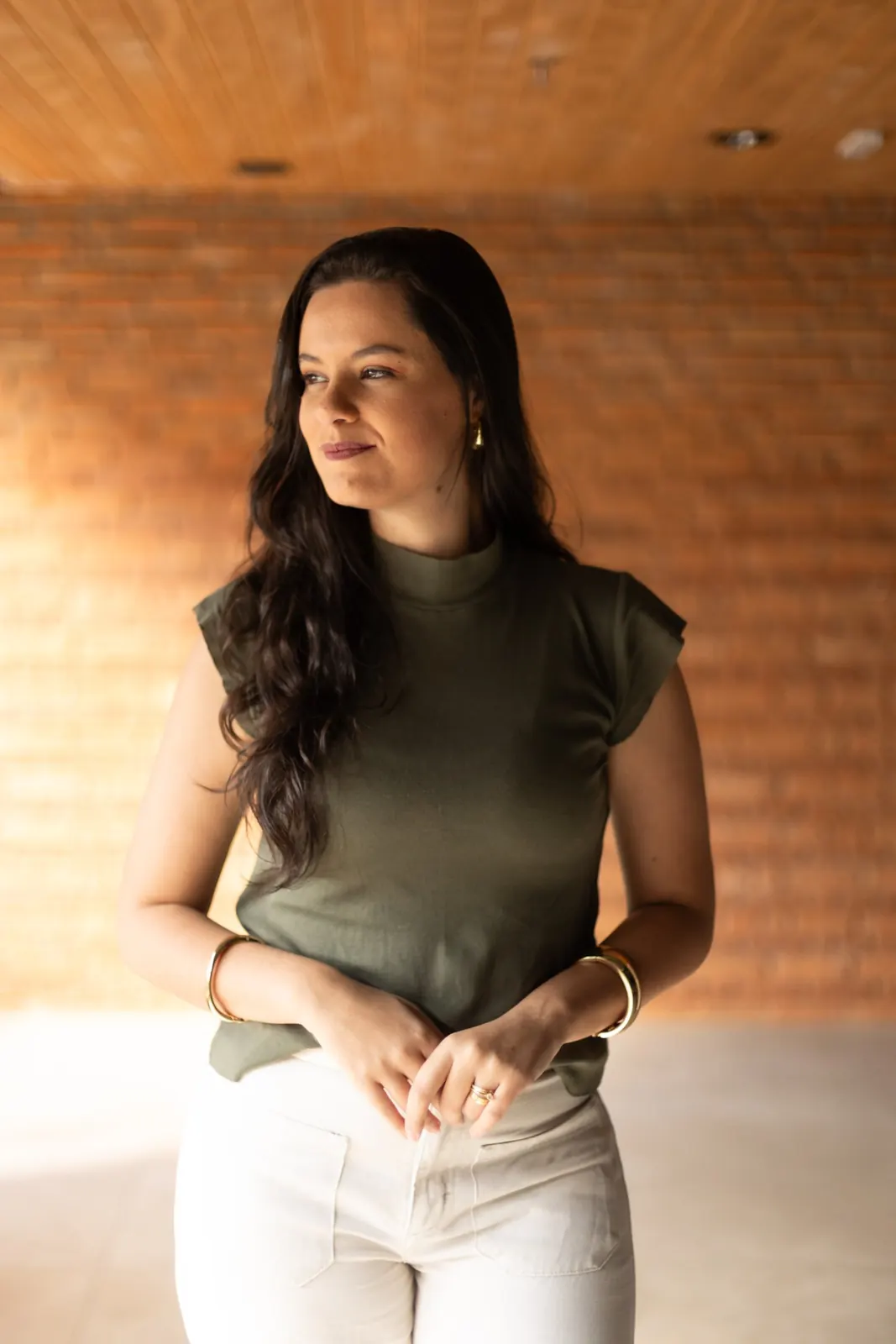 Raquel diante de uma parede de tijolos, olhando para o lado com um leve sorriso
